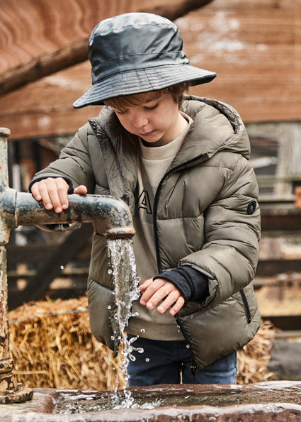 Child in a green jacket and hat playing with water from a faucet outdoors.
