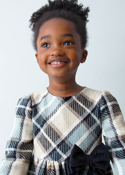 Young girl wearing a plaid dress with a bow, smiling against a plain background