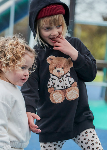 Two children, one wearing a black hoodie with a teddy bear design, standing outdoors.
