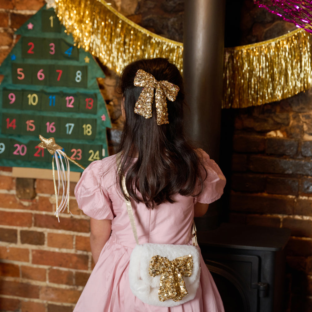 A young girl with long dark hair wearing a pink dress and a gold sequined bow in her hair, seen from behind. She is wearing a white faux-fur crossbody bag with a large gold sequin bow on the front, standing in front of a brick fireplace with gold tinsel garlands.