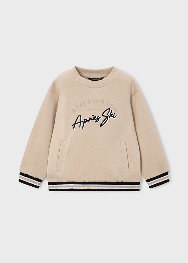 A beige crewneck sweatshirt with black embroidered text reading 'Real Snow Brand' and 'Après Ski' on the front, featuring black and white striped ribbed cuffs and hem.