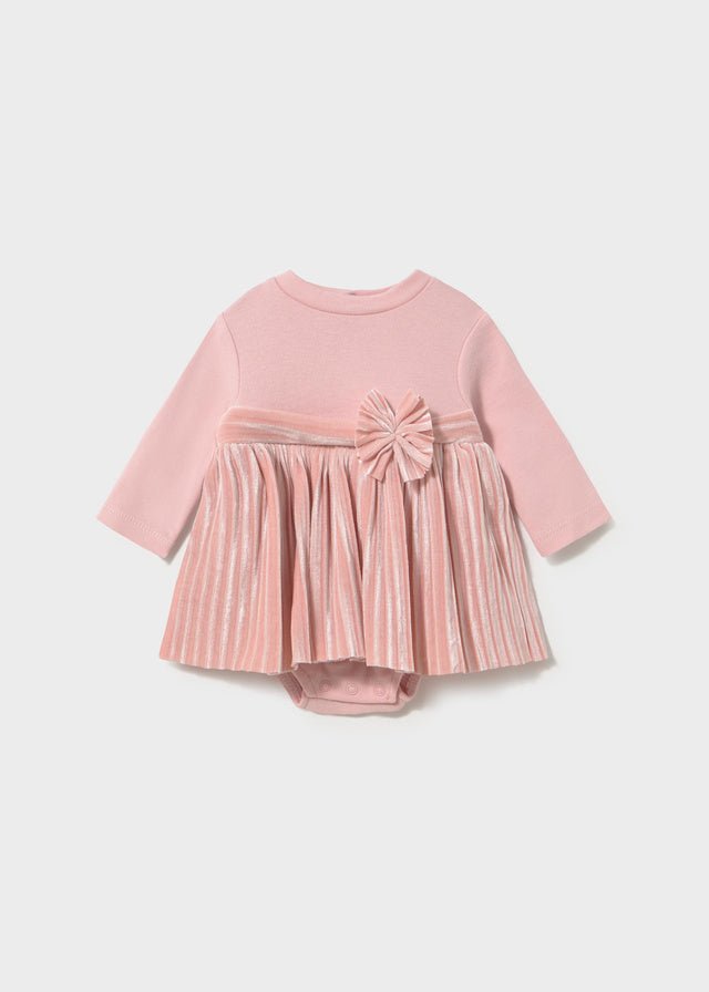 A baby girl's long-sleeve romper in dusty pink, featuring a ribbed knit top, a pleated velvet skirt, and a matching fabric bow on the chest, displayed against a light gray background.