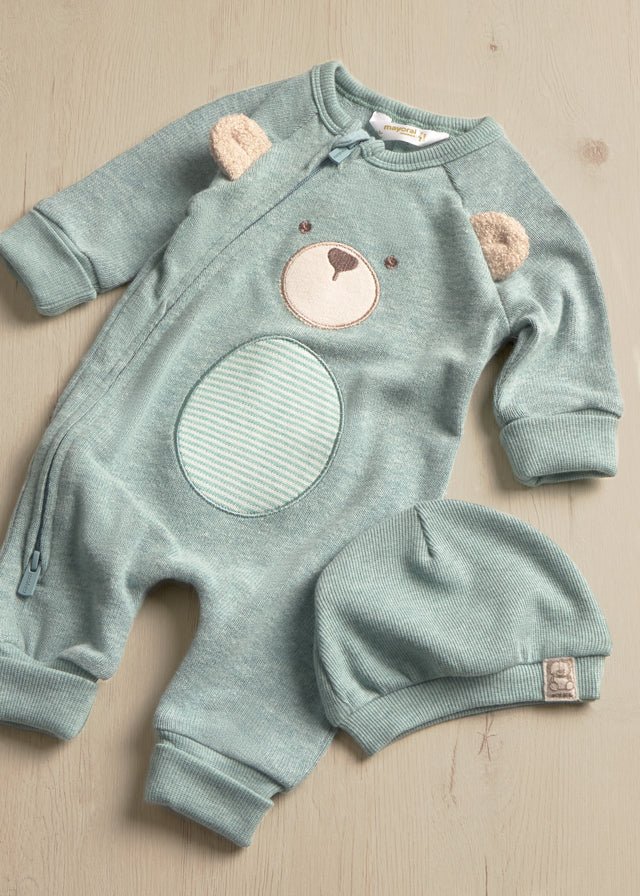 A baby's long-sleeve footless onesie in a dusty sage green, featuring a bear face applique on the chest, a striped bear belly patch, and two small bear ear patches. A matching knit bonnet is placed next to it on a light wood background.