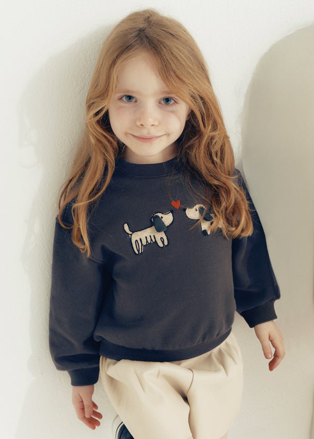 Young girl wearing a navy blue sweatshirt with dog designs against a white wall.