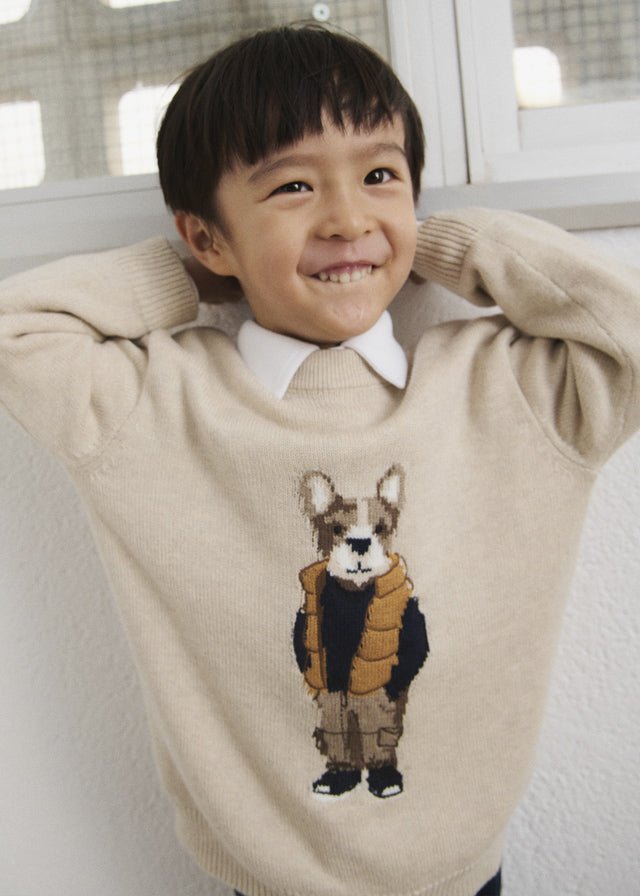 A young boy smiling and posing indoors, wearing a beige knit sweater with a detailed dog character graphic on the front.