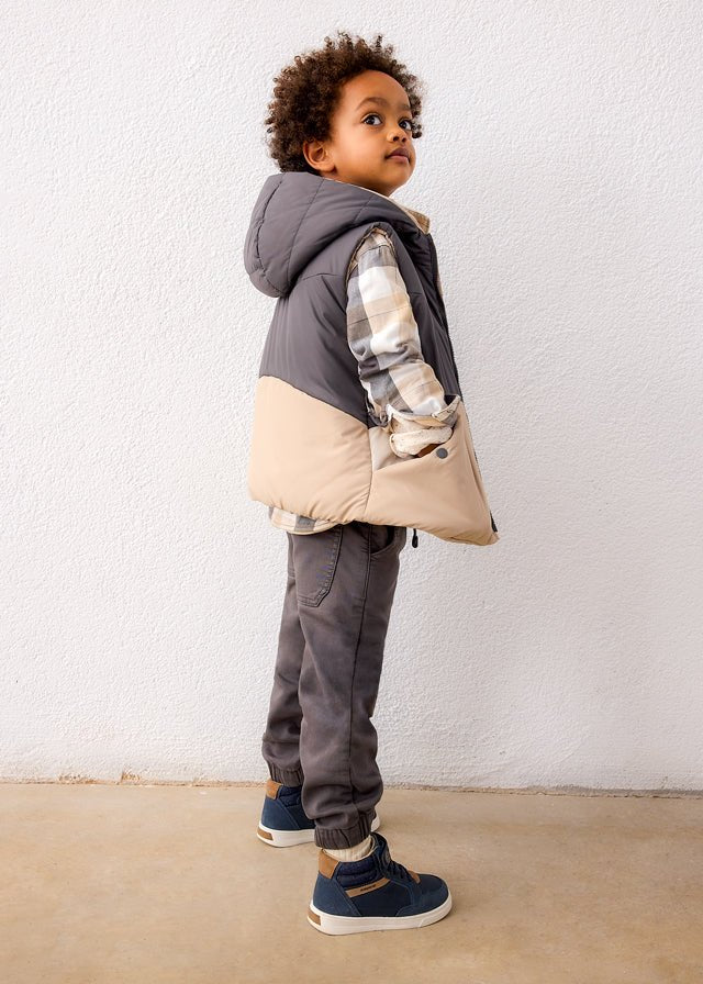 Child wearing a gray and beige jacket with a hood, standing against a white wall.