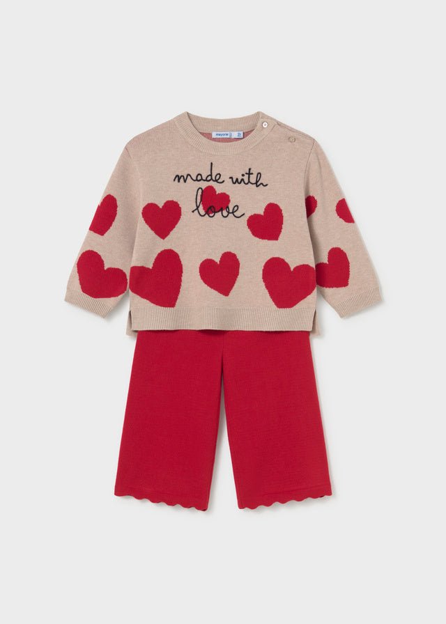 A children's knit two-piece set featuring a beige long-sleeve top with red hearts and 'made with love' text, paired with solid red pants, shown on a light gray background.