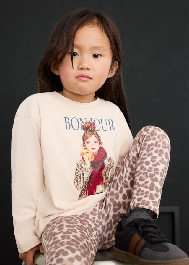 Child wearing a 'Bonjour' sweatshirt with a graphic design and leopard print pants against a dark background.