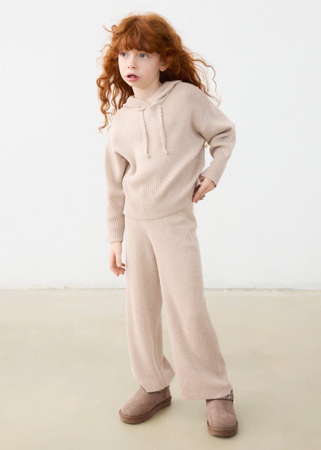 A young girl with curly red hair stands in a studio wearing a matching beige knit hoodie and wide-leg pants set, paired with brown suede boots against a plain light background.