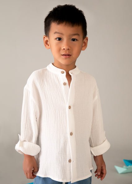 Boy wearing a mayoral white long sleeve cheesecloth style button up shirt, with rolled sleeves and grandad style collar.