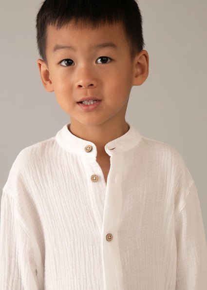 Boy wearing a mayoral white long sleeve cheesecloth style button up shirt, with rolled sleeves and grandad style collar.