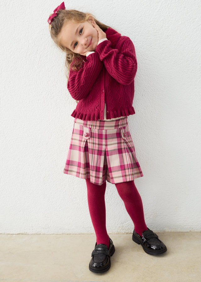 Young girl wearing a red sweater, plaid shorts, and tights against a white background