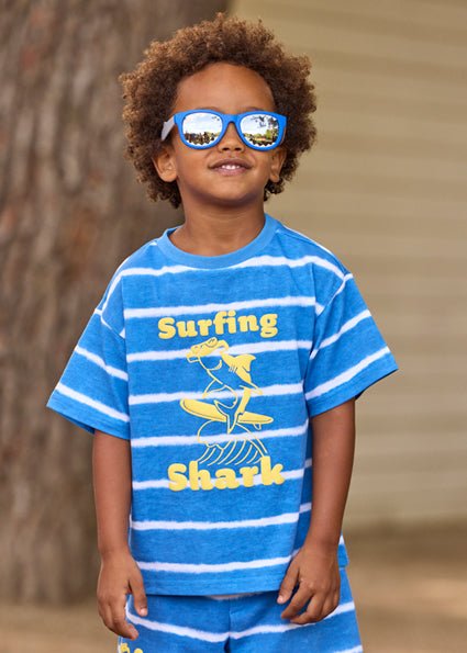 Child wearing a blue and white striped shirt with 'Surfing Shark' design, standing against a natural background.