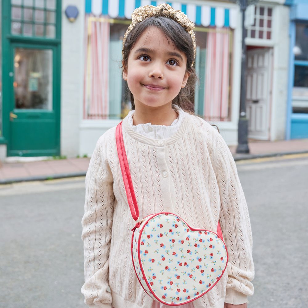 Young girl wearing a cream sweater and heart-shaped bag with a cherry pattern, standing on a street.