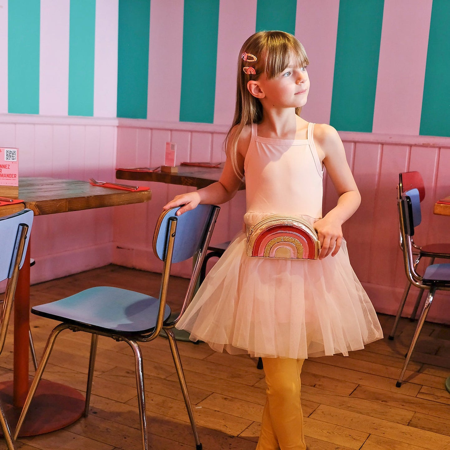 Young girl in a pink dress holding a red and gold bag in a room with striped walls.