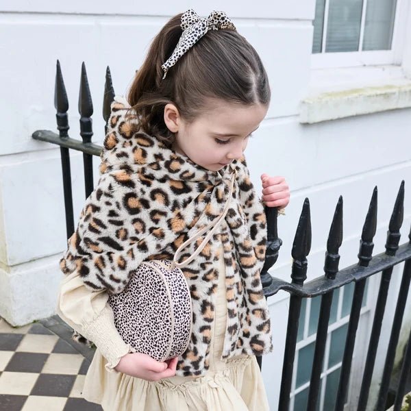 child wearing leaopard print hair scarf bobble, bag and hooded ponch, next to railings and a chequered floor