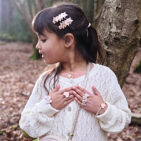 A young girl with dark hair in a ponytail stands outdoors against a tree trunk, wearing a cream cable-knit cardigan over a pink top. She is accessorized with a pink and white star hair clips.
