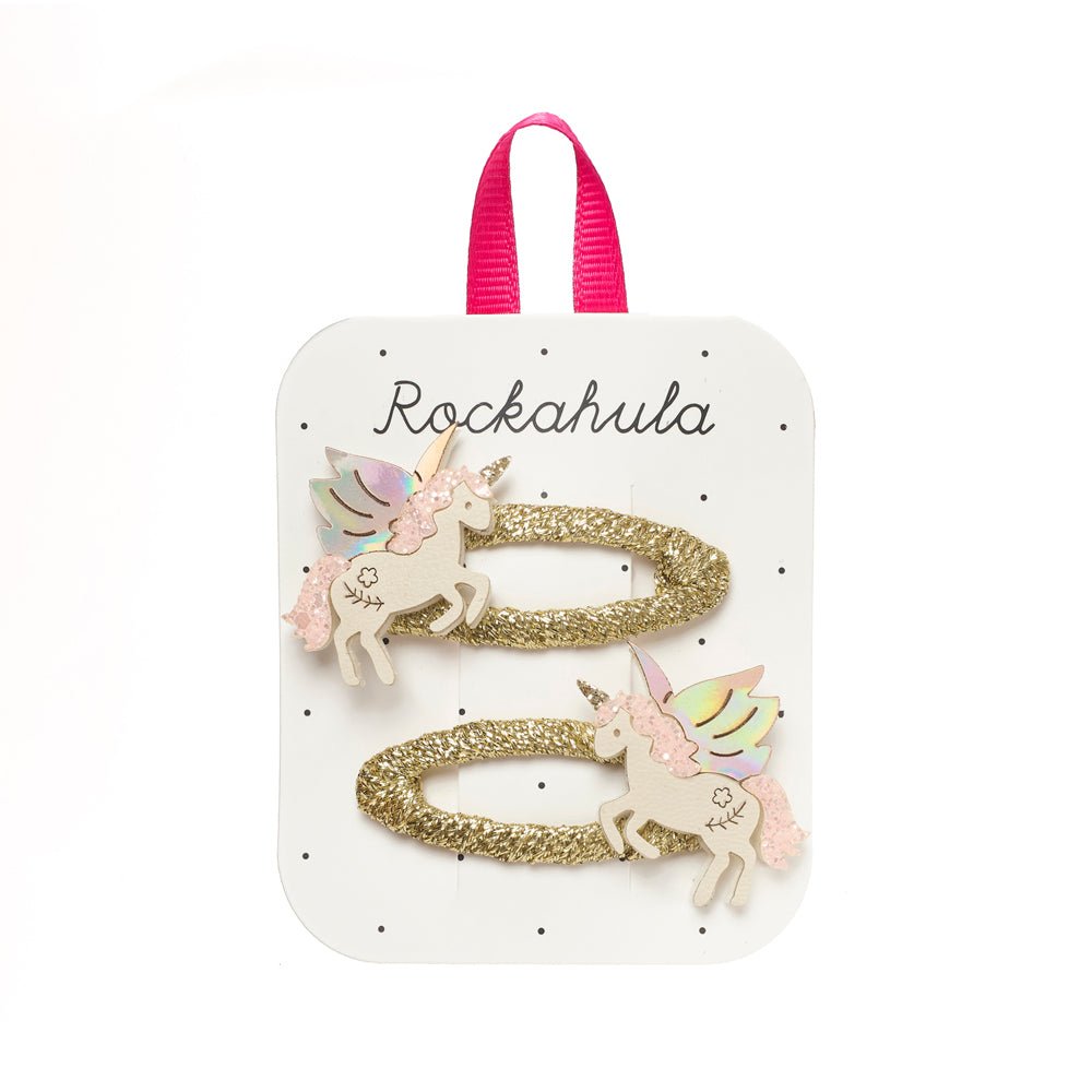 A pair of gold glitter hair clips featuring cute unicorn designs, presented on a white display card with a pink ribbon loop. The card is branded with the name 'Rockahula'.